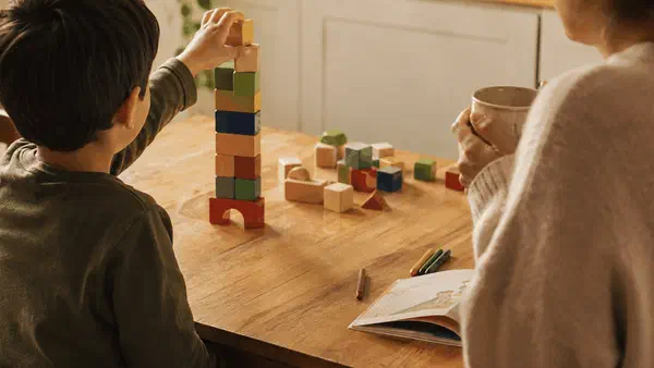 Young child stacking wooden blocks at a kitchen table while a parent watches nearby