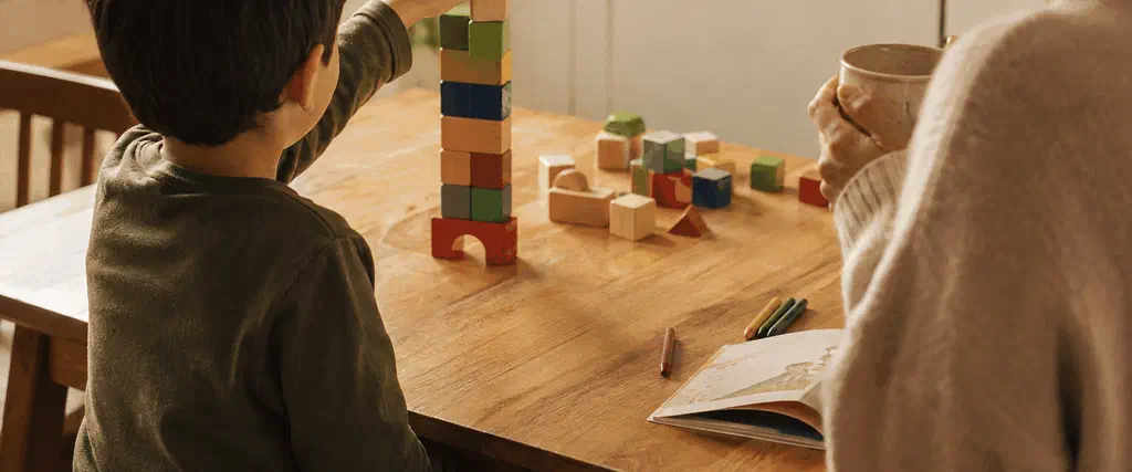 Young child stacking wooden blocks at a kitchen table while a parent watches nearby